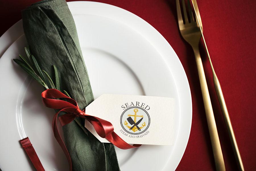 Close up view on setting plate with tag and golden cutlery. Christmas table setting with Seared logo overlay